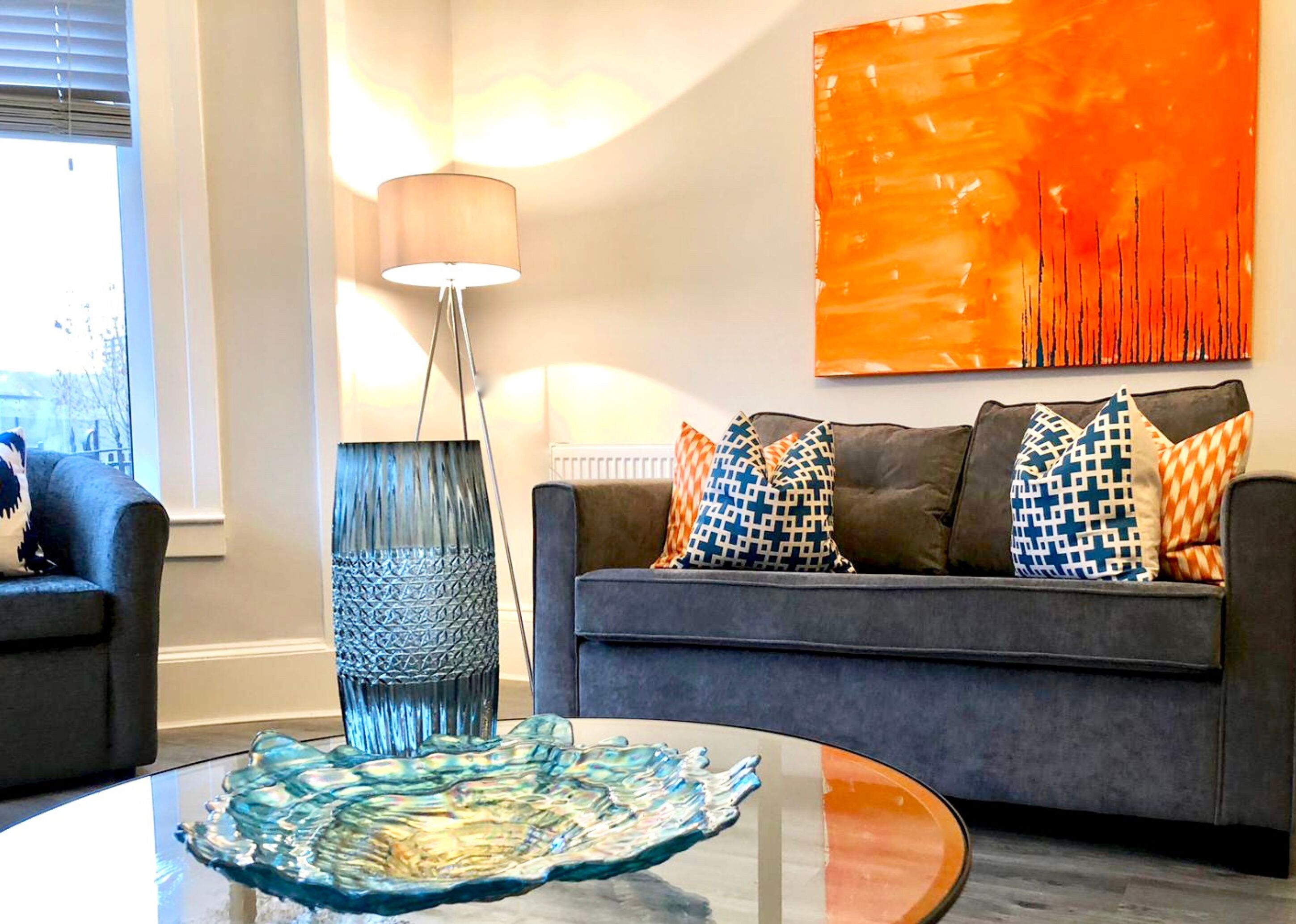 Een moderne woonkamer met een grijze bank, gestreepte kussens, een blauwe vaas en een glazen decoratiemiddenstuk op de tafel.