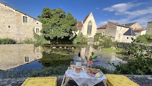 A stone table with a bottle of wine, a glass, and a flower arrangement set on a stone ledge by a calm waterway. - Les Bruyères (Verteuil-sur-Charente)