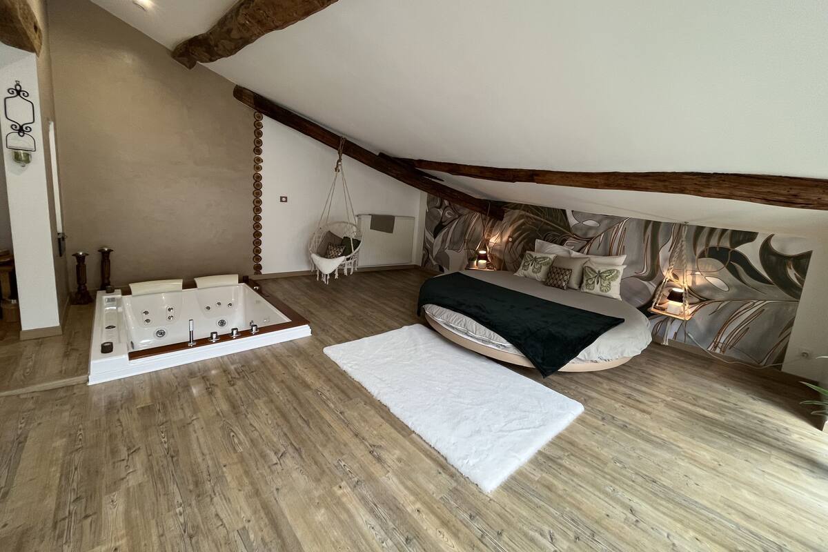 Een slaapkamer met een grote houten vloer, een bed met veel kussens, een hangstoel en een bad.