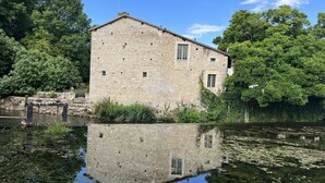 A stone building with a small window and a chimney, surrounded by trees and a calm body of water reflecting its structure. - Les Bruyères (Verteuil-sur-Charente)