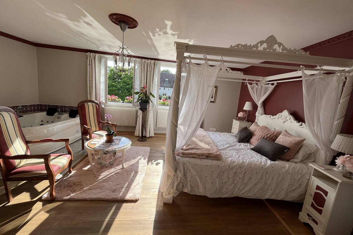Een slaapkamer met een hemelbed, een zithoek met een stoel en een tafel, een badkuip, en uitzicht op groen buiten.