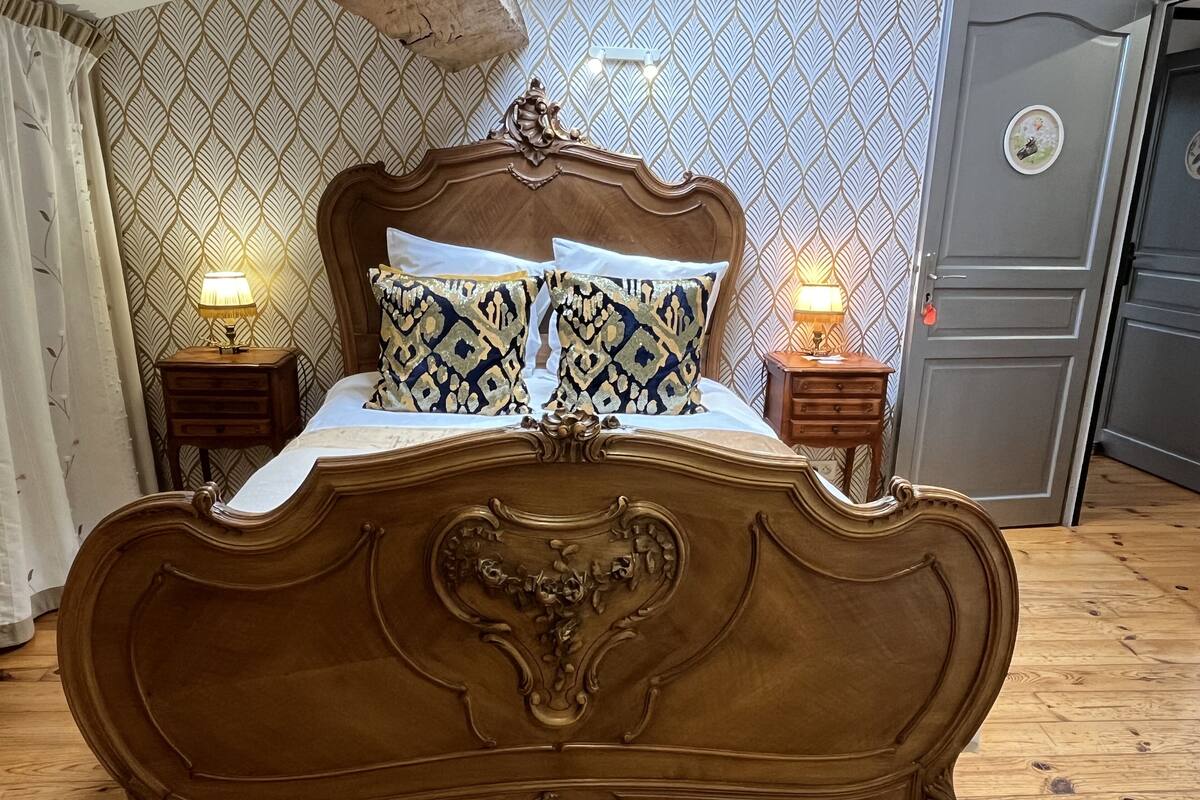 Een slaapkamer met een groot, versierd bed, twee nachtkastjes met lampen, behang met een patroon en een houten vloer.