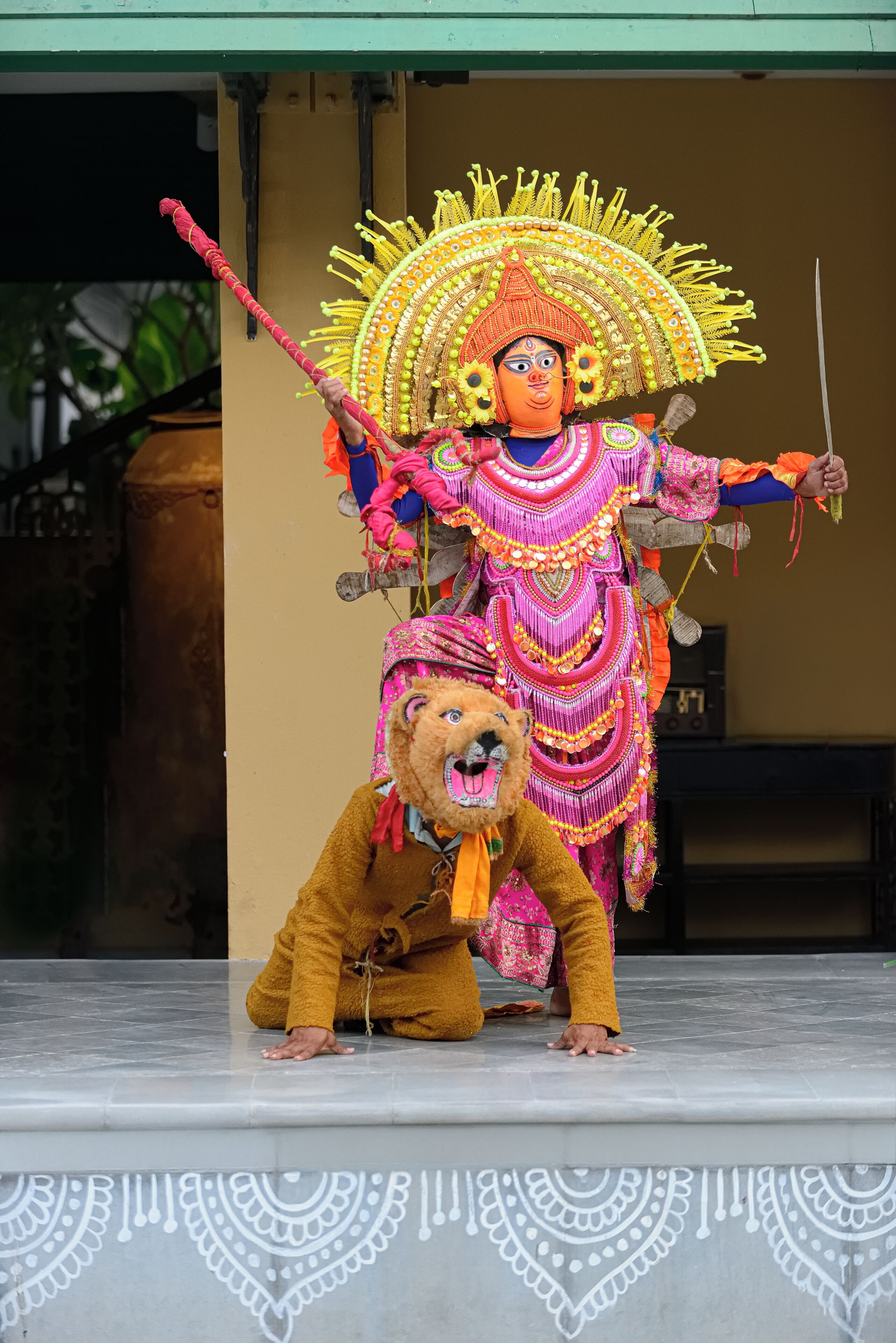 A person in a colorful costume with a large headdress and a prop stick, standing on a stage with a lion mask.