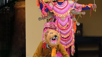 A person in a colorful costume with a large headdress and a prop stick, standing on a stage with a lion mask.