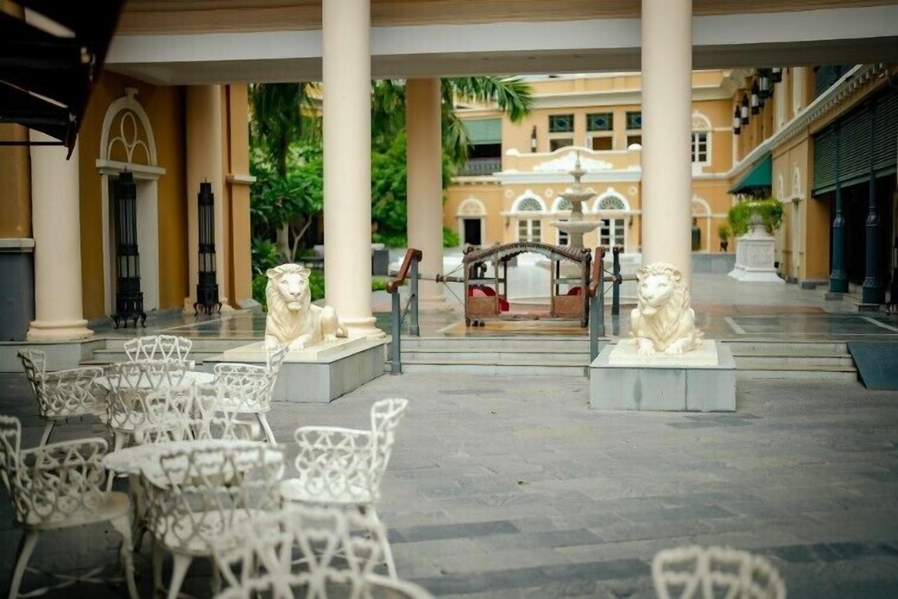 Une cour avec des chaises et des tables en métal blanc, deux statues de lions et une fontaine en arrière-plan.