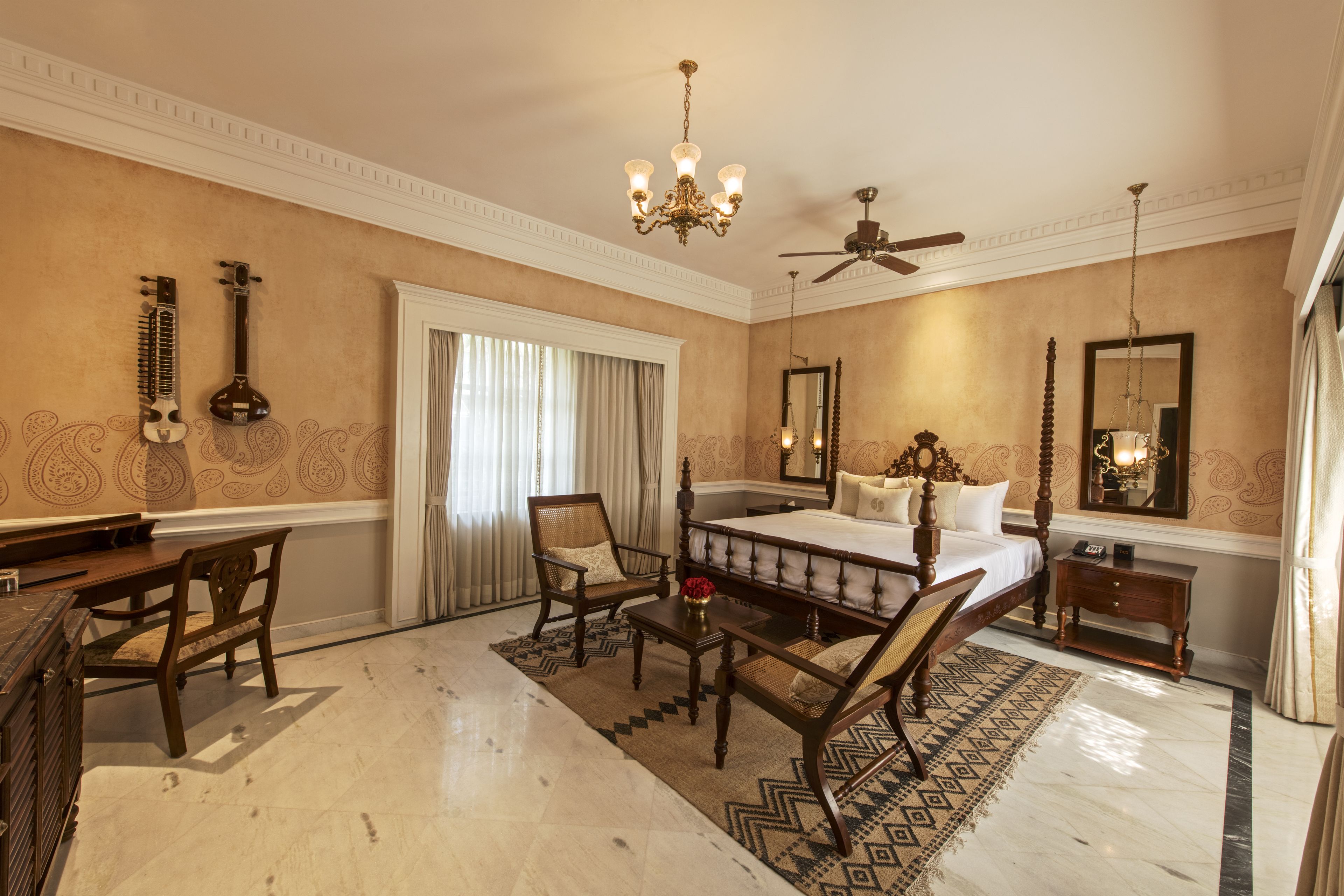 A spacious bedroom with a large bed, a wooden desk, two chairs, a chandelier, a ceiling fan, and a large mirror.