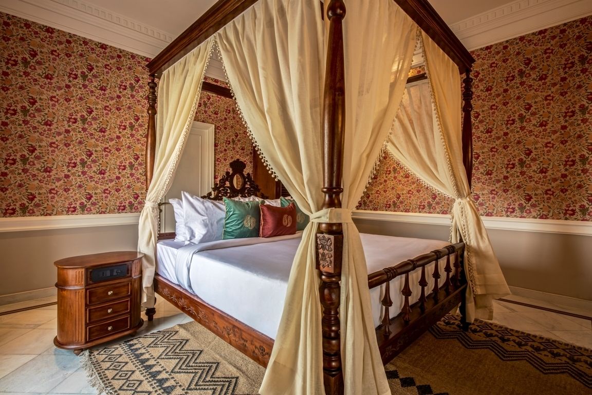 A four-poster canopy bed with a wooden frame and white drapes, a nightstand with drawers, and a patterned rug.