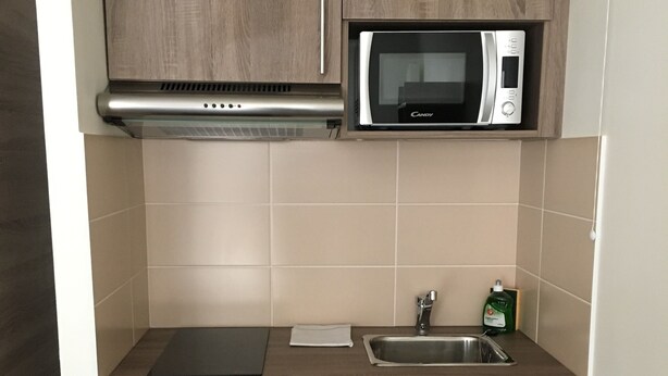 A compact kitchen with wooden cabinets, a microwave, a built-in oven, a sink, and a refrigerator. - KOSY Appart’Hôtels – Art’ & Facts (Nancy)