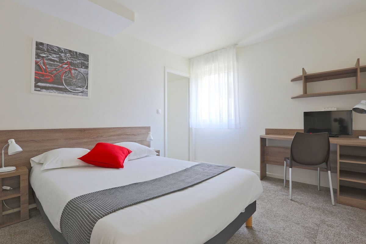 A hotel room with a bed, a desk with a computer, a chair, a window with curtains, and a painting on the wall.