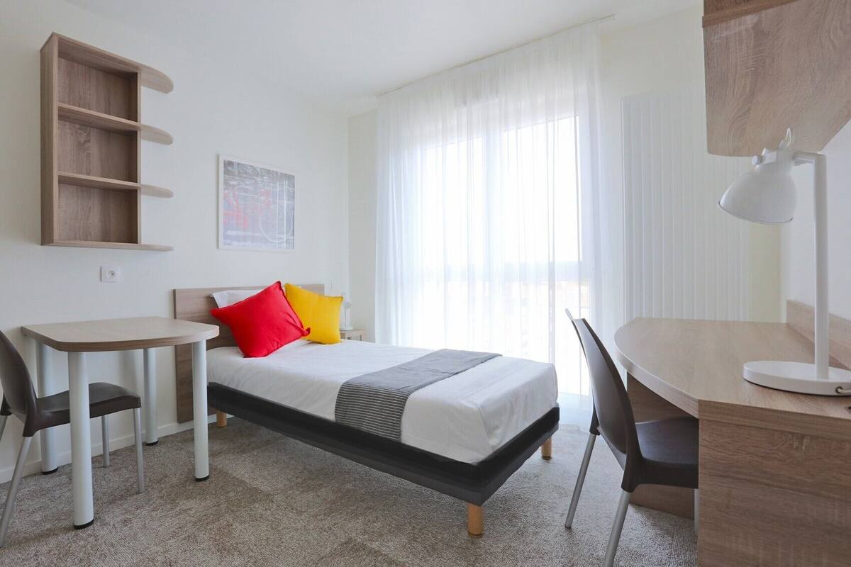 A hotel room with a bed, a desk, a chair, a small table, and a shelf.