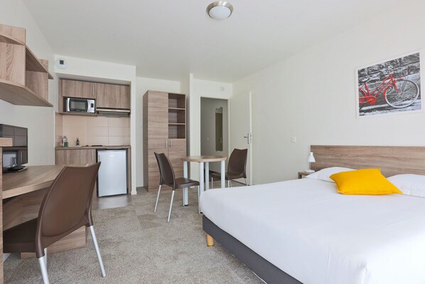 A hotel room with a bed, a desk, a chair, a microwave, and a small kitchenette. - KOSY Appart’Hôtels – Art’ & Facts (Nancy)