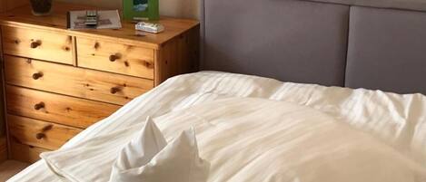 Un letto rifatto con cuscino con la scritta "Teotonia Hotel", un comò di legno con un vaso di fiori e un quadro appeso al muro.