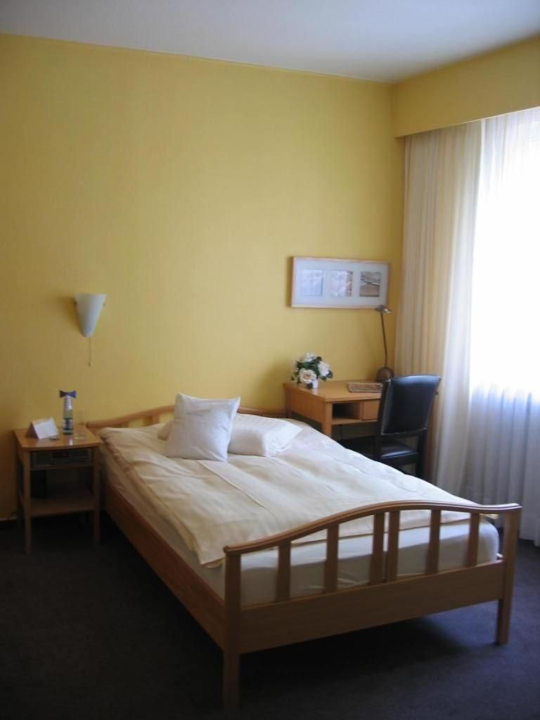 A hotel room with a single bed, a desk, a chair, a lamp, and a window with curtains.