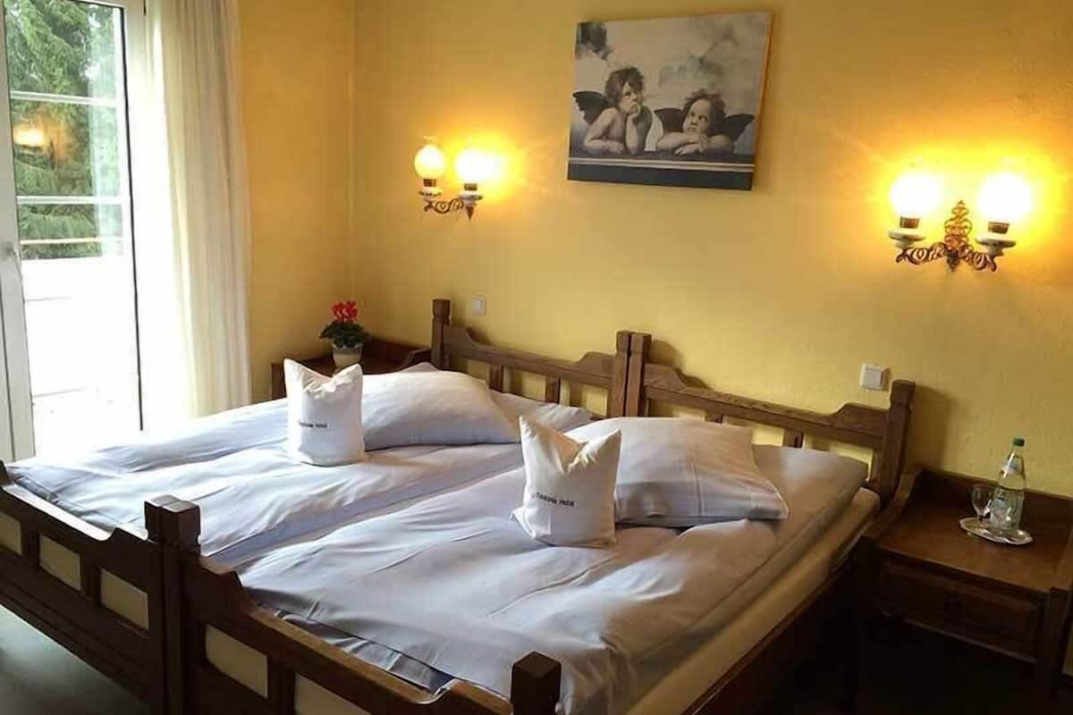 A double bed with white linens, two wall-mounted lamps, a painting above the bed, and a nightstand with a bottle and glass.