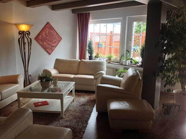 Un salon avec un canapé beige, une table basse en verre et un lampadaire. Il y a une fenêtre avec des rideaux et une vue sur un jardin.
