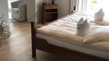 A neatly made bed with a white pillow and a white throw blanket, a wooden nightstand with a phone and a small plant, a painting of flowers on the wall, and a view of the outdoors through a window.