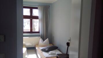 A small, cozy room with a bed, a chair, a window with curtains, and a ceiling fan.