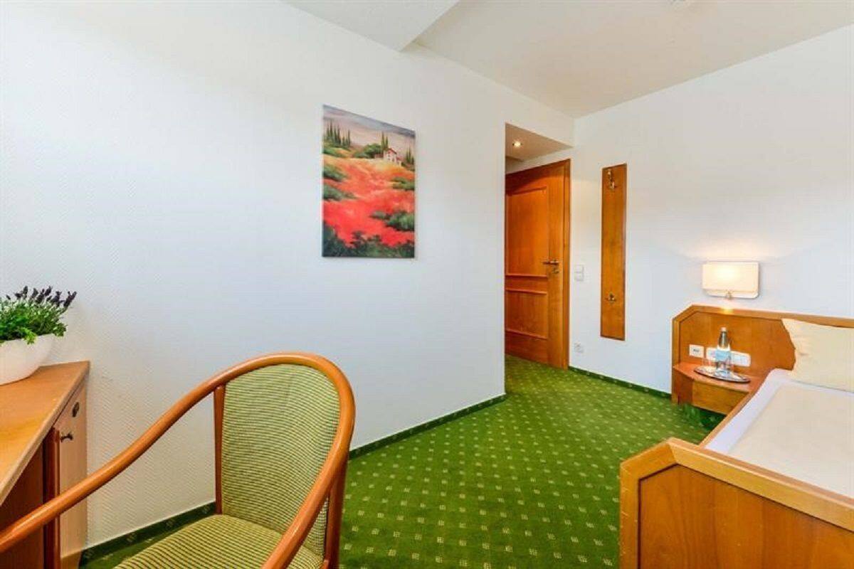 A hotel room with a green carpet, a wooden chair, a bed with a wooden headboard, a bedside table with a lamp, and a painting on the wall.