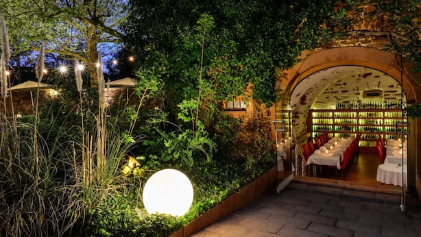 An outdoor dining area with tables and chairs, surrounded by lush greenery and a stone archway leading to a bar area. - Hotel Jauch's Löwen (March)