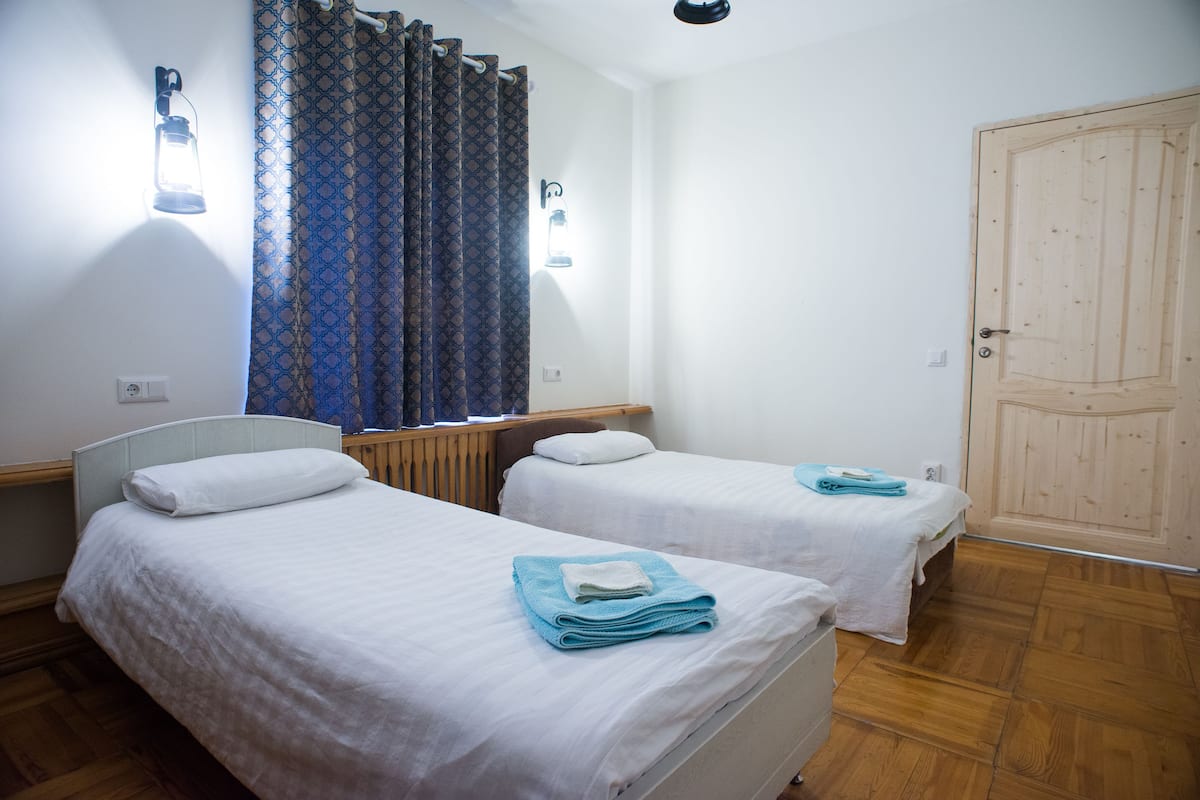Une chambre avec deux lits simples, chacun doté d’un matelas blanc et d’oreillers assortis. On y trouve une porte en bois et une fenêtre ornée de rideaux à motifs.
