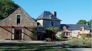 Ein Steinhaus mit großem Rundbogenportal und einem kleineren Fenster, umgeben von einer gepflegten Rasenfläche und einem Metallzaun.