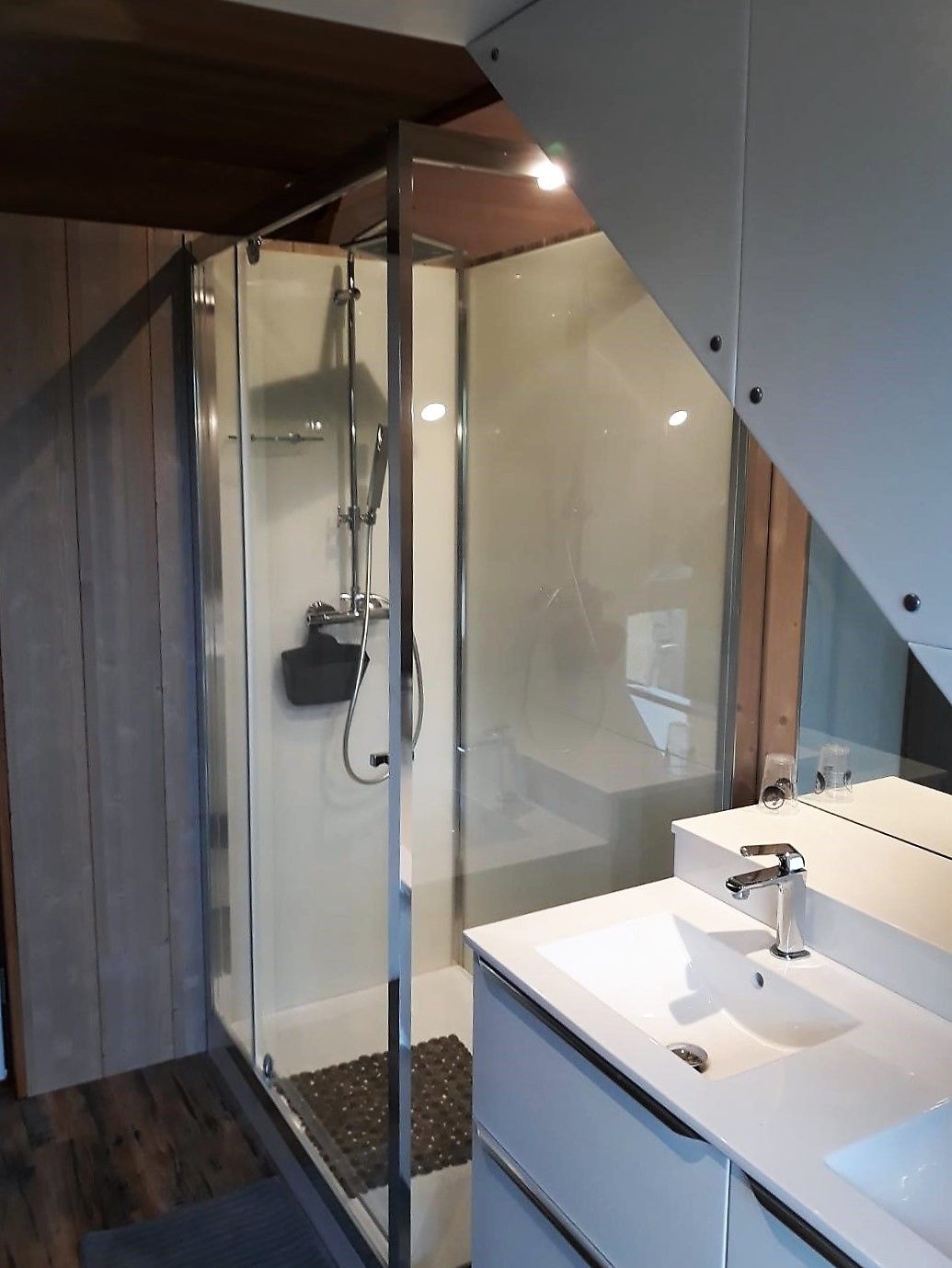 Un baño moderno con cabina de ducha de vidrio, tocador blanco con espejo y suelo de madera.