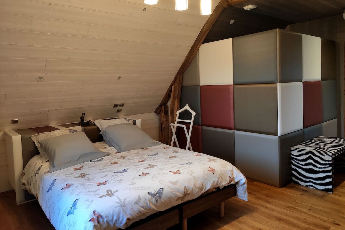 Un dormitori tipus loft amb sostre inclinat, bigues de fusta, un llit amb roba de llit floral, una panxa i una paret acolchida i colorida.