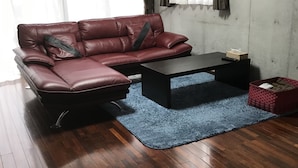 A living room with a brown leather sofa, a black coffee table, a blue rug, wooden flooring, and a wall-mounted shelf with baskets. - Biimata house (Nago)