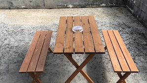 A wooden picnic table with benches, set on a concrete surface with a concrete wall featuring circular cutouts in the background. - Biimata house (Nago)