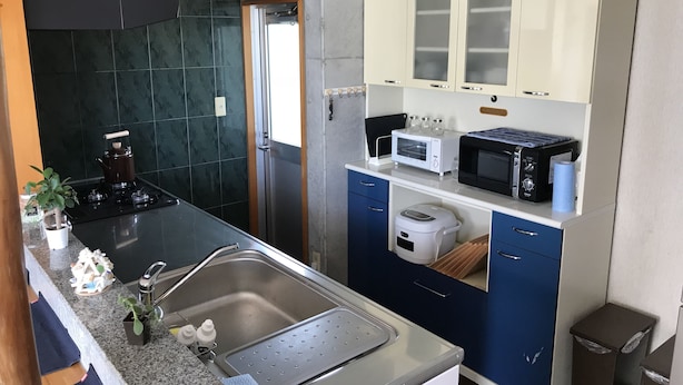 A compact kitchen with a stainless steel sink, a microwave, a toaster, and a small oven. There are cabinets above the counter and a trash bin to the right. - Biimata house (Nago)