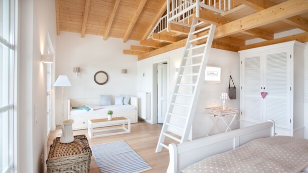 Una habitación luminosa y amplia con zona superior, vigas de madera, cama blanca, sofá, escalera y armario.