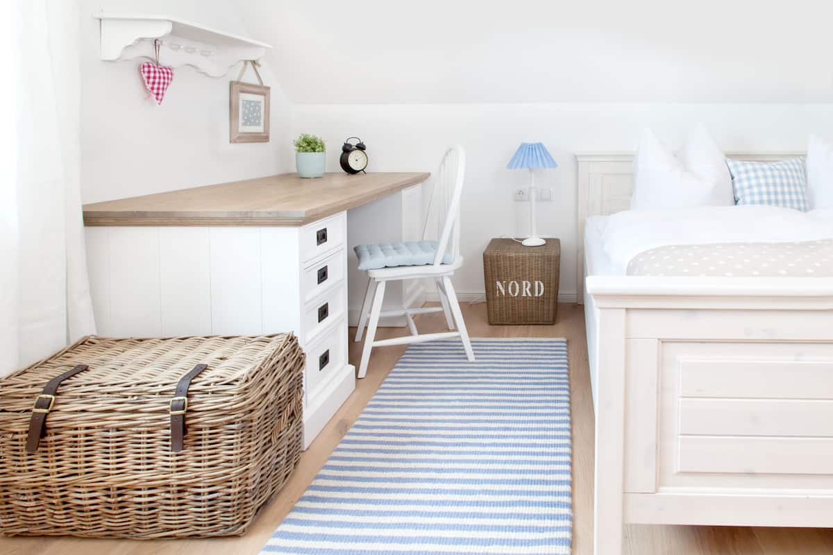 A bedroom with a bed, a desk, a chair, a wicker basket, and a blue-striped rug.
