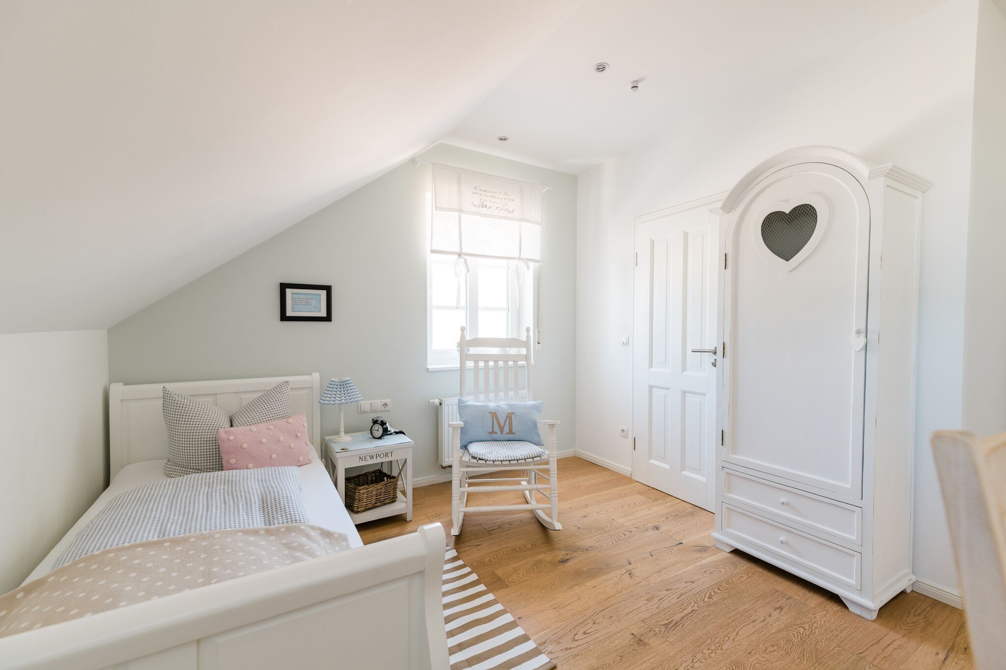 A bedroom with a sloped ceiling, a bed with a striped bedspread, a rocking chair, a white wardrobe with a heart-shaped window, and a small bedside table with a lamp.