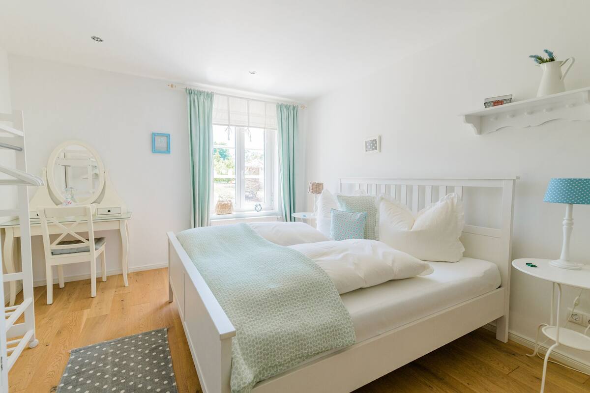 A bedroom with a bed, a desk, a chair, a small table, and a shelf with decorative items.