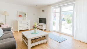 Una sala moderna con un sofá, una mesa de centro blanca con un jarrón, un televisor y un amplio ventanal corredera que da a un espacio exterior.
