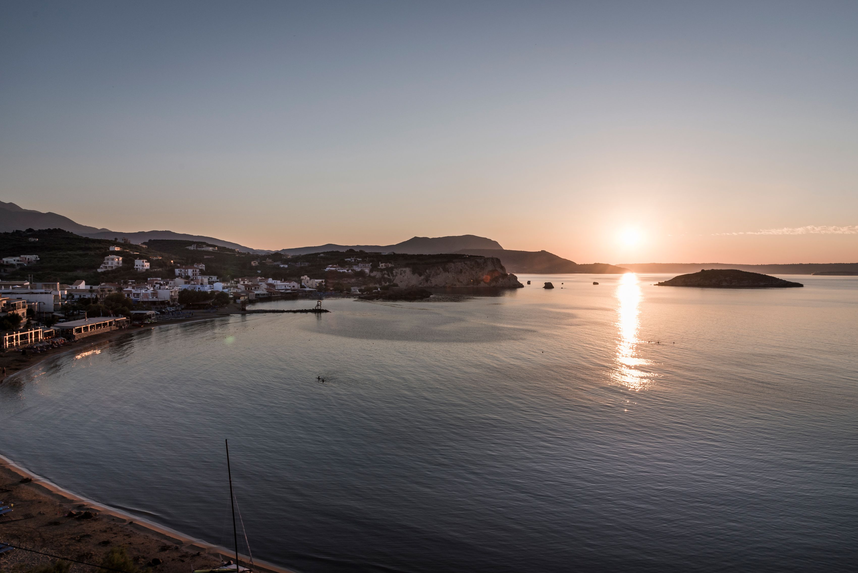 Una località costiera con edifici lungo il lungomare, acque calme e un tramonto sullo sfondo.