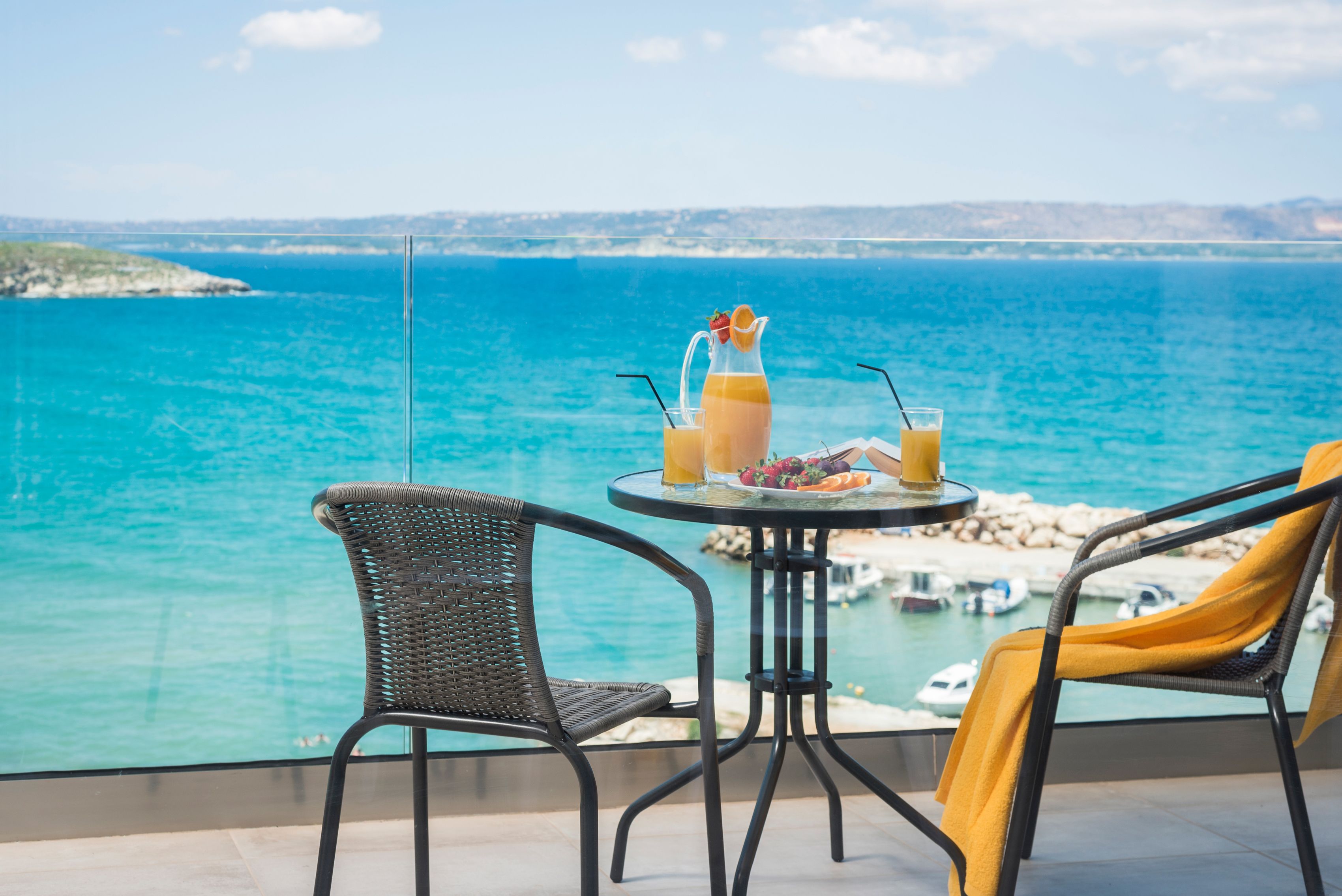 A balcony with a view of the sea, two wicker chairs, a round table with drinks and fruit, and a yellow towel.