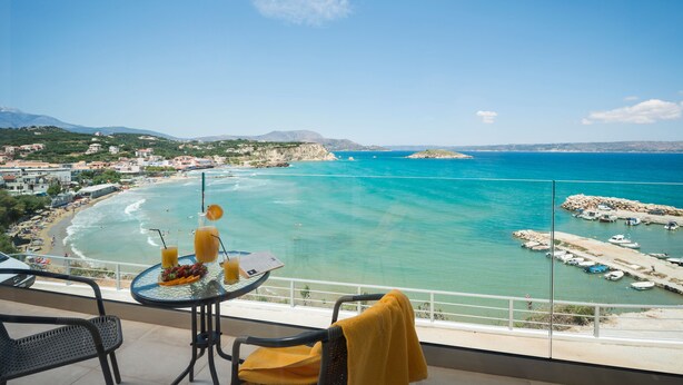 A balcony with a table and chairs overlooking a beach and marina. - Panorama Plus (Apokoronas)