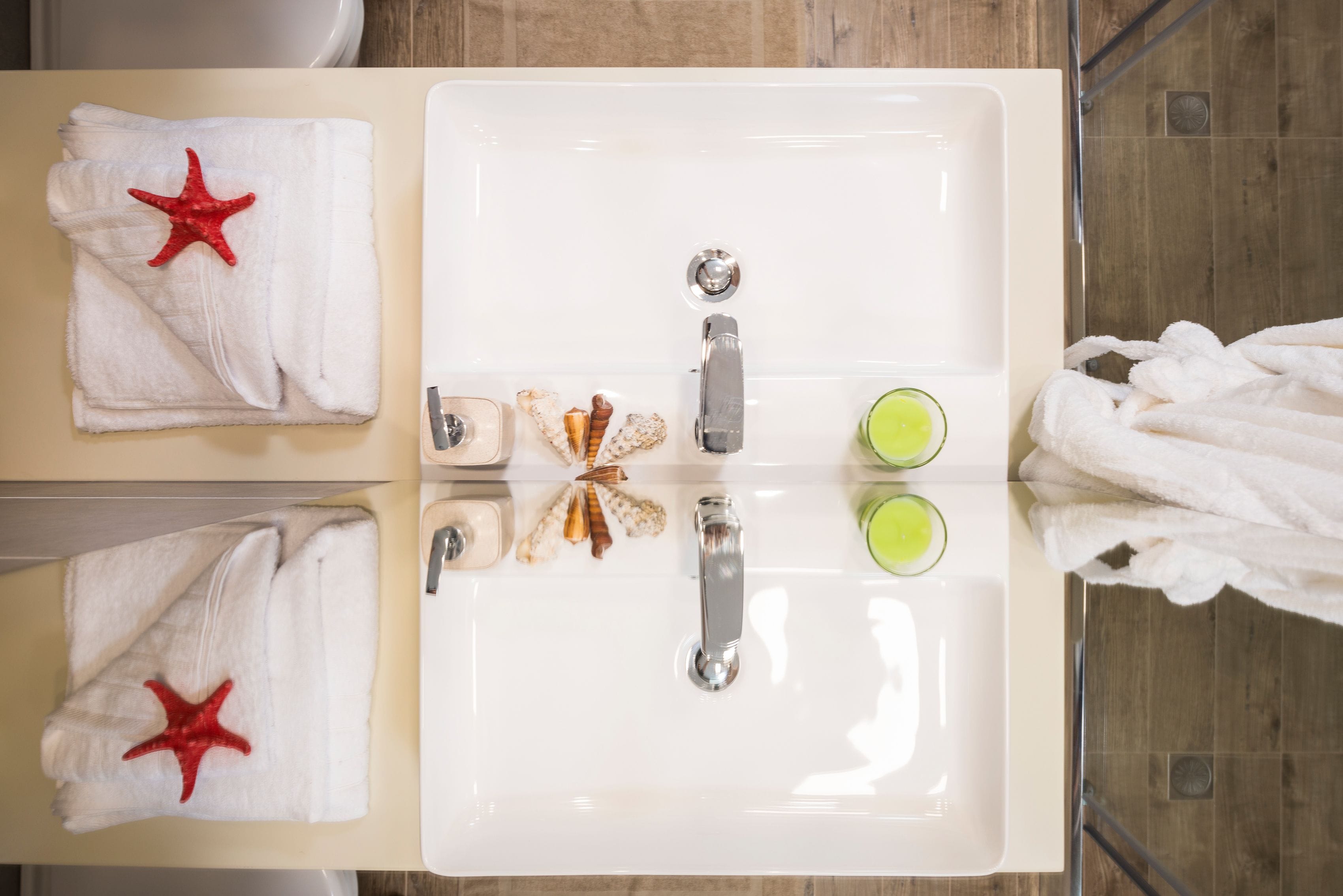 Une salle de bain avec un lavabo blanc, deux robinets et un miroir. On y trouve des serviettes ornées d’étoiles de mer rouges et un verre contenant un liquide vert posé sur le lavabo.