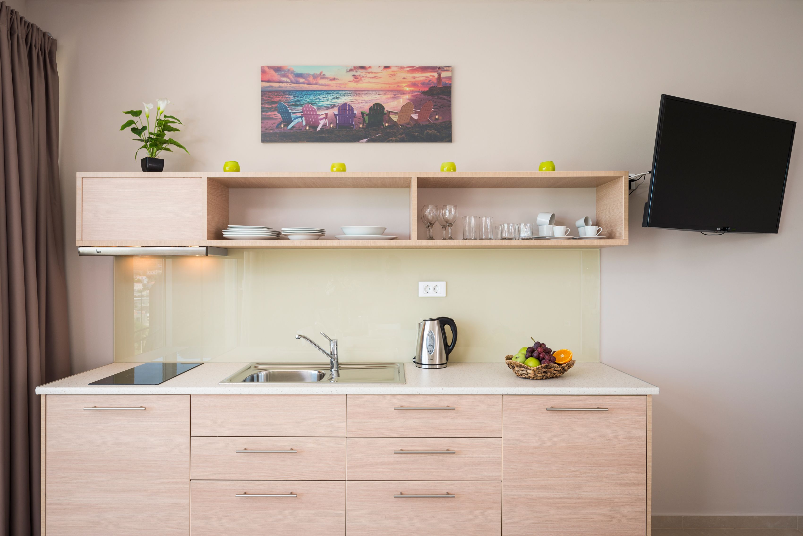 Une cuisine moderne équipée d’une cuisinière intégrée, d’un évier et d’un plan de travail. Une étagère au-dessus de l’évier est utilisée pour ranger les assiettes et les verres, et un panier de fruits est posé sur le plan de travail.