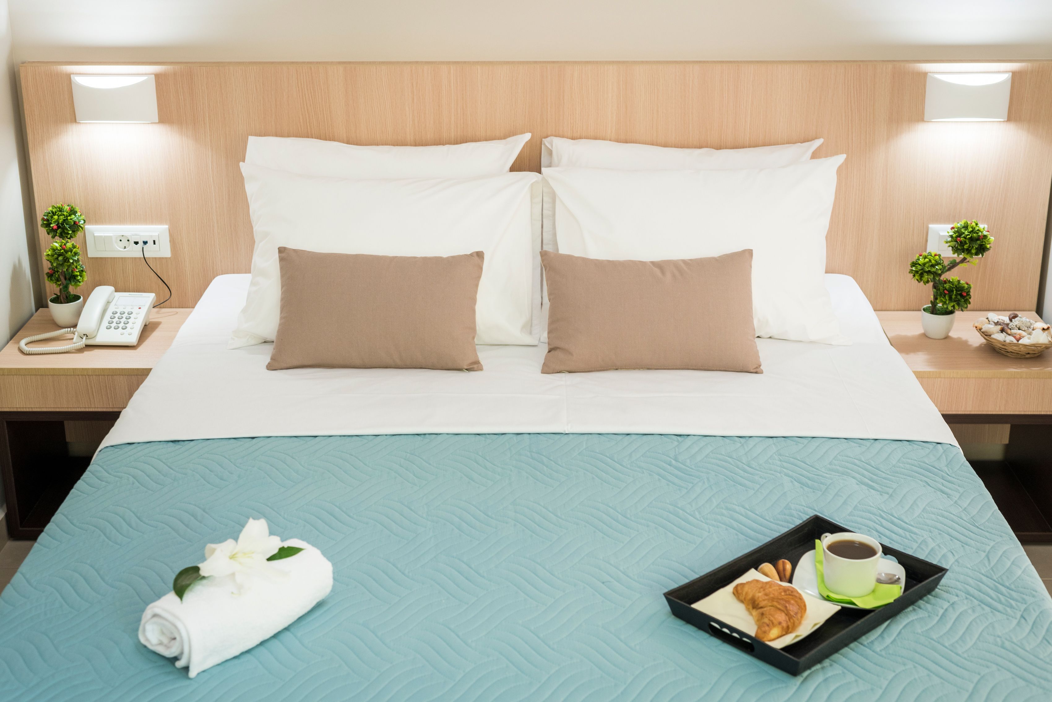 A hotel room with a bed, two bedside tables, a nightstand, a phone, a tray with croissants and coffee, and a rolled towel with flowers.