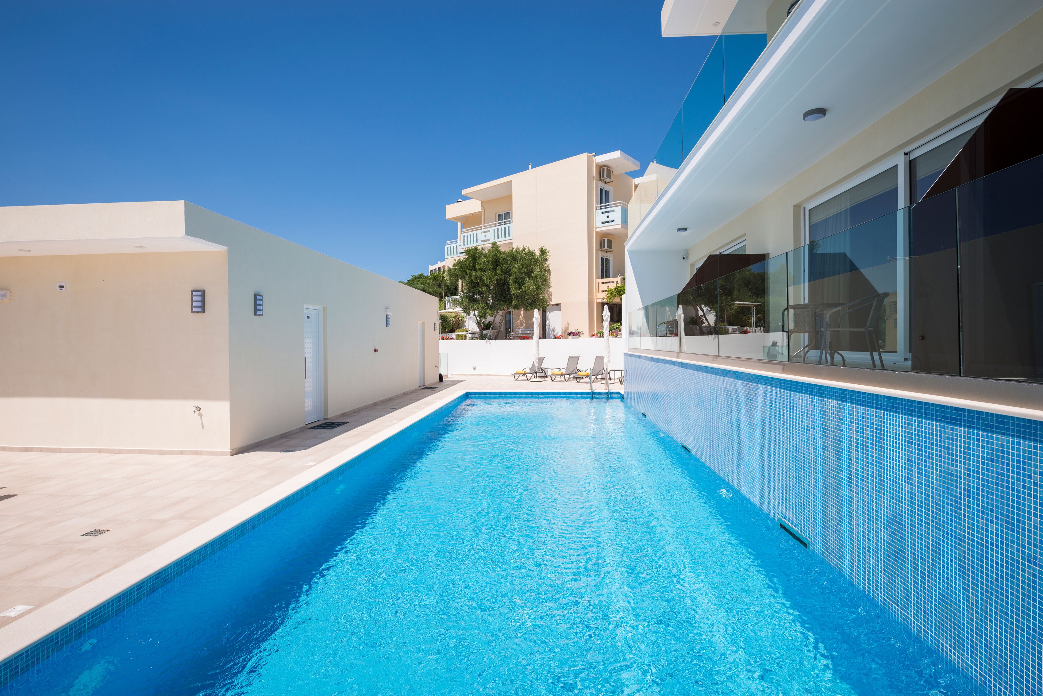 A swimming pool with clear blue water, surrounded by a modern building with balconies and a tiled deck area.