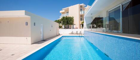 A swimming pool with clear blue water, surrounded by a modern building with balconies and a tiled deck area.