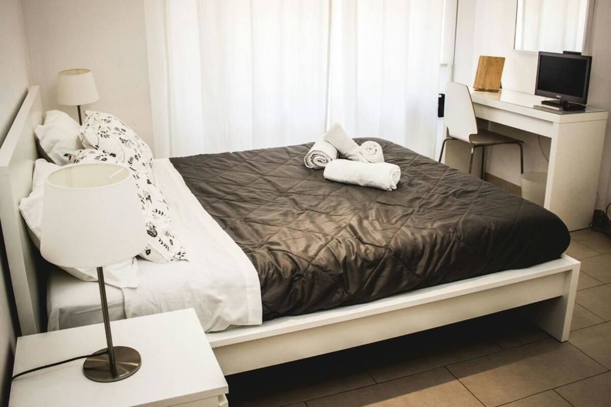 A neatly made bed with a dark quilt, a bedside lamp, and a desk with a computer monitor.