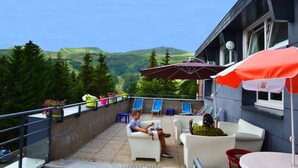 Um terraço com mesas ao ar livre, vista para colinas verdes e um edifício com fachada escura.