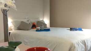 Quarto de hotel com cama, mesa de cabeceira com abajur, caneca de café e um quadro na parede.