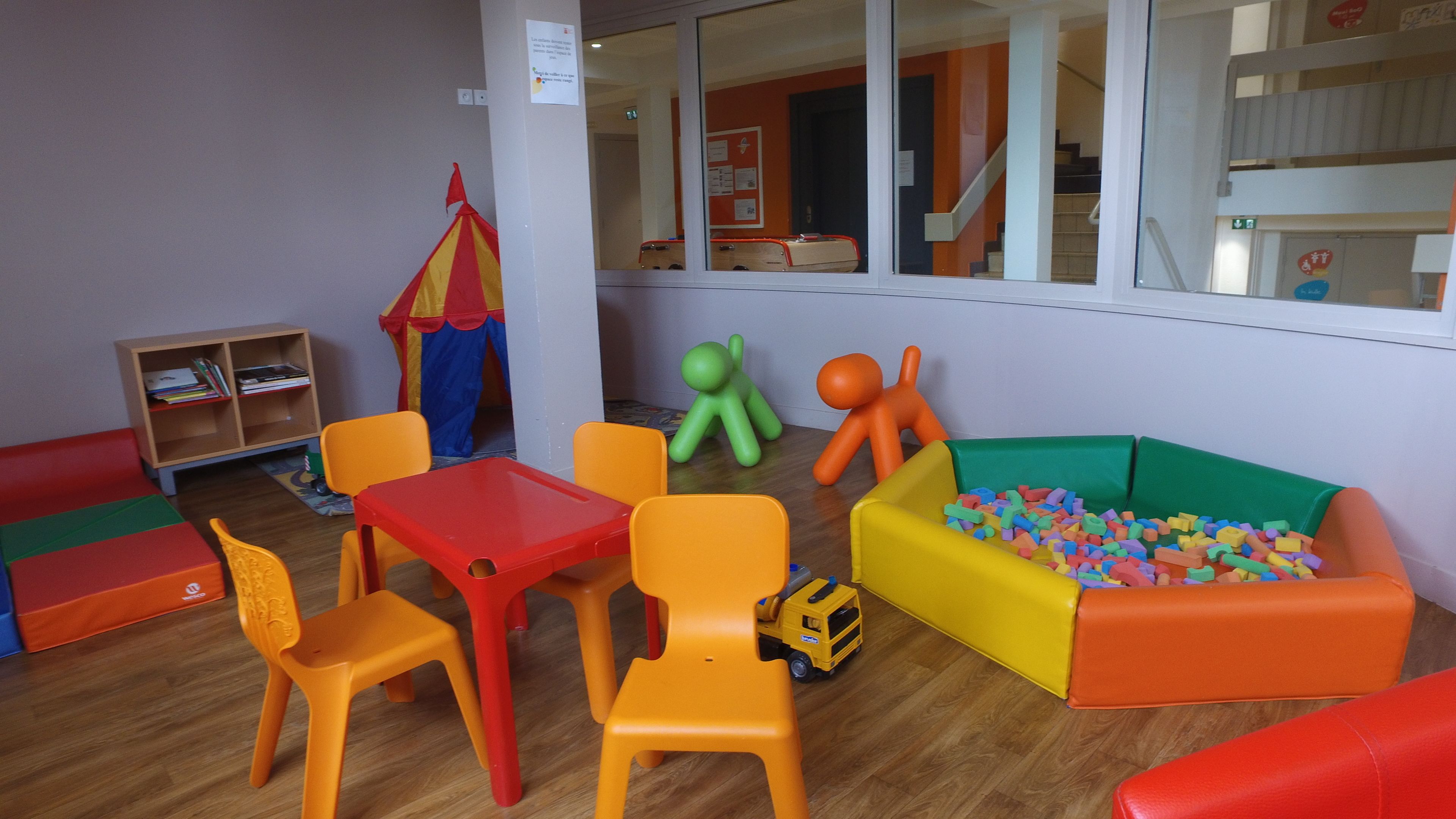Un espace de jeux pour enfants avec des meubles colorés, notamment une table rouge, des chaises orange et un parc pour enfants avec des blocs de mousse.