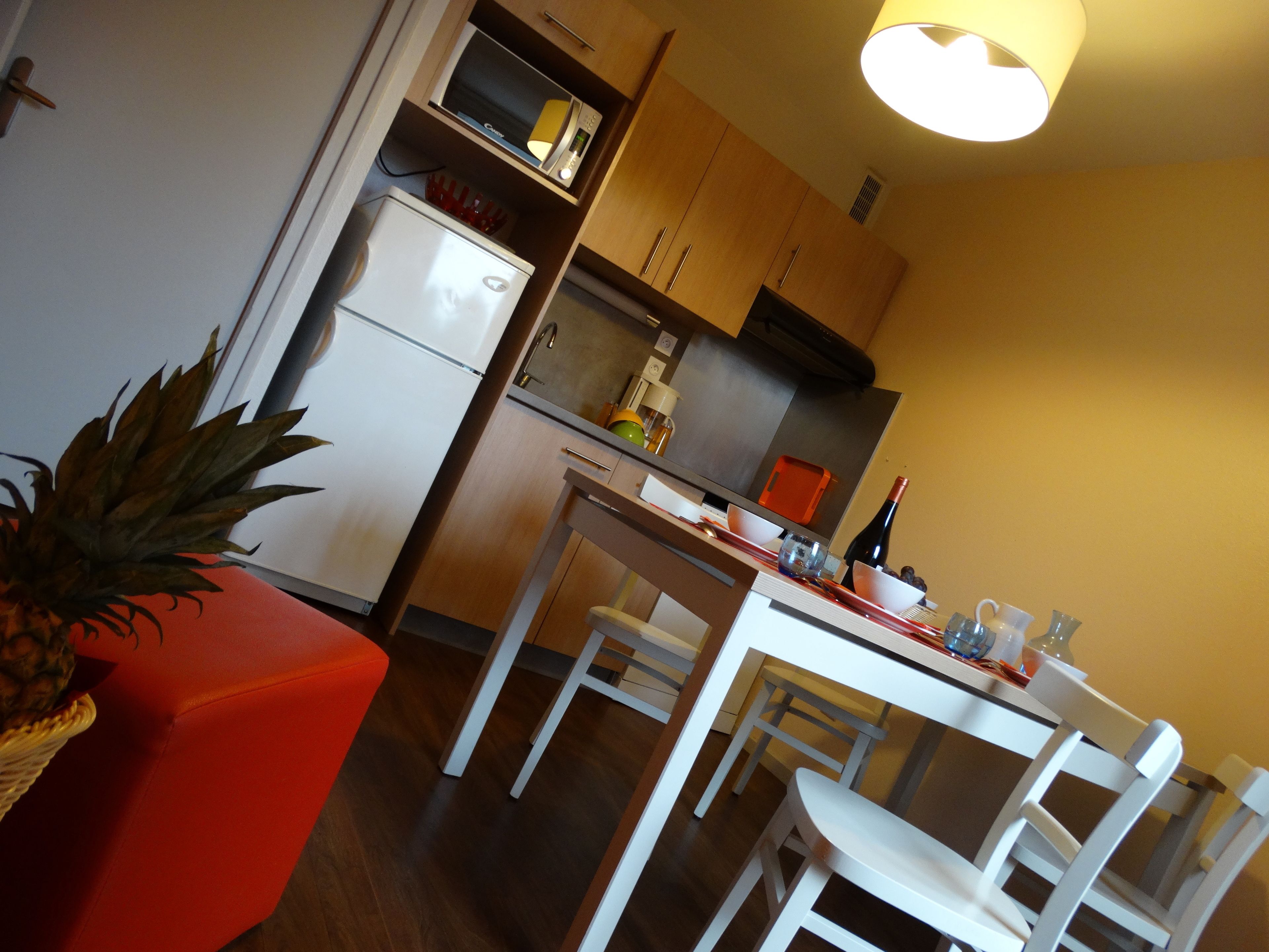 A compact kitchen with a refrigerator, microwave, and stove. There is a dining table with chairs and a small red ottoman.