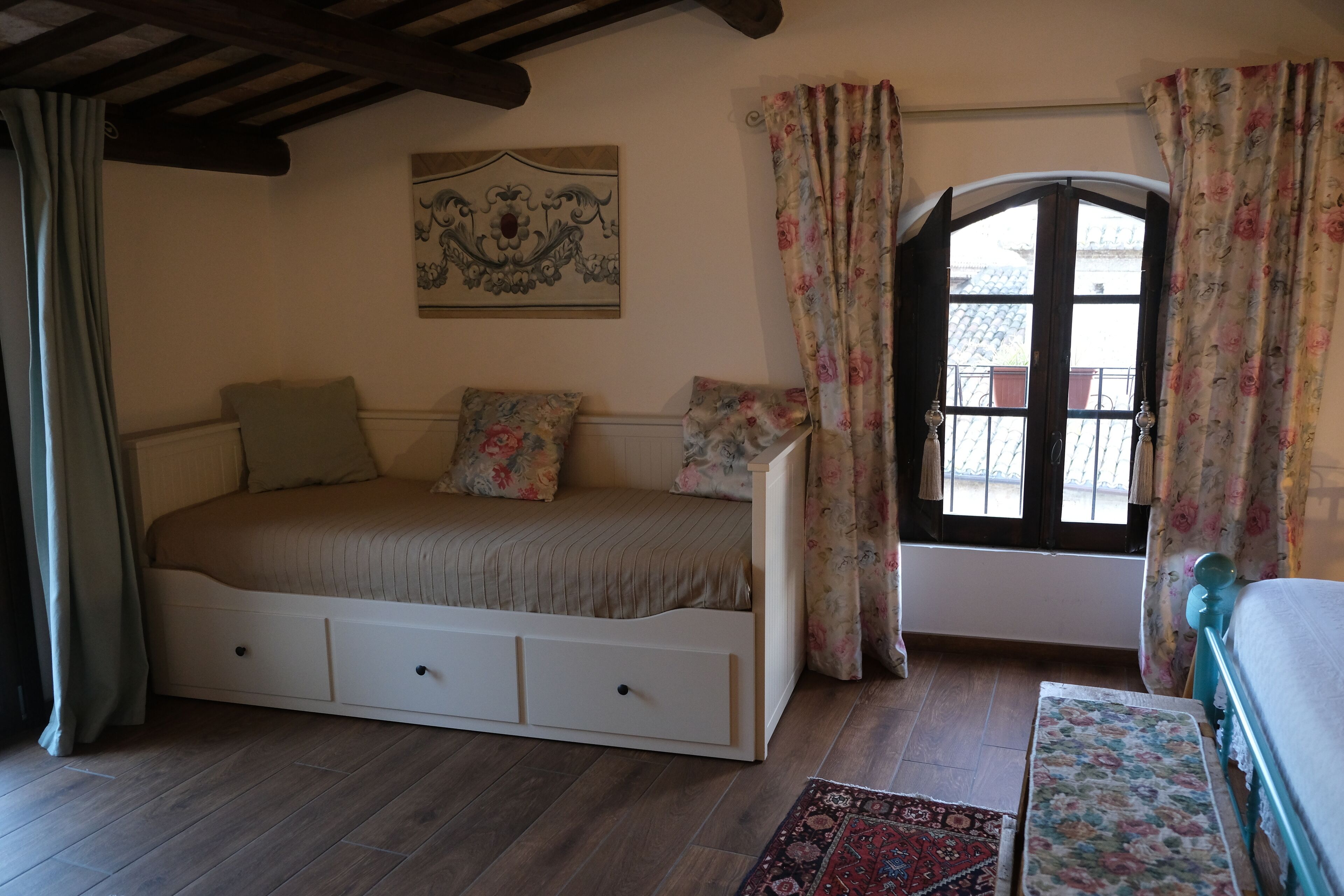 A bedroom with a bed, a window with curtains, a decorative wall piece, and a wooden floor.