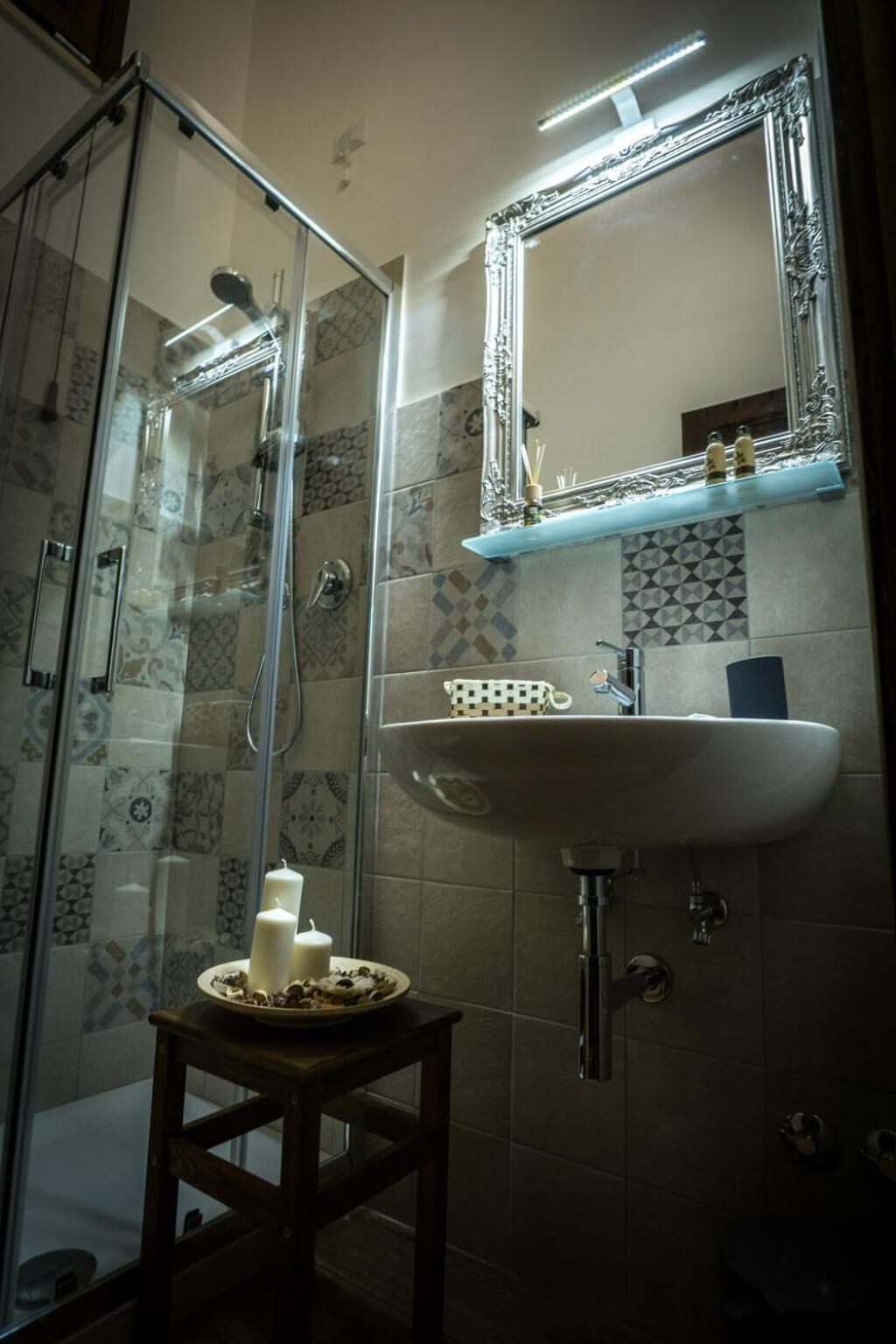 Une salle de bain avec une douche à parois de verre, un lavabo avec un miroir au-dessus et un petit tabouret sur lequel sont posées des bougies.